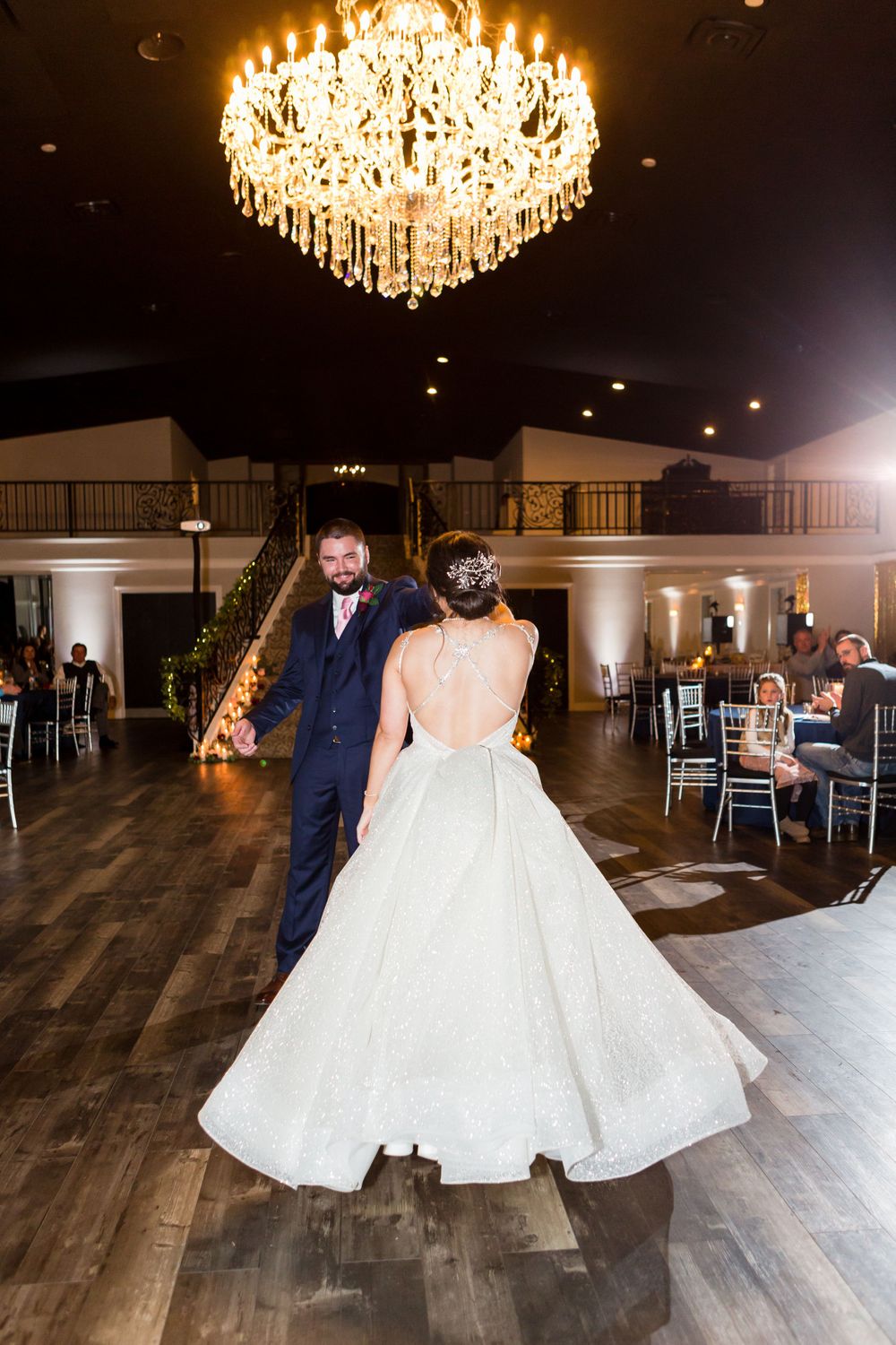 A couple dances in an elegant ballroom under a crystal chandelier with hardwood floors and guest seating.