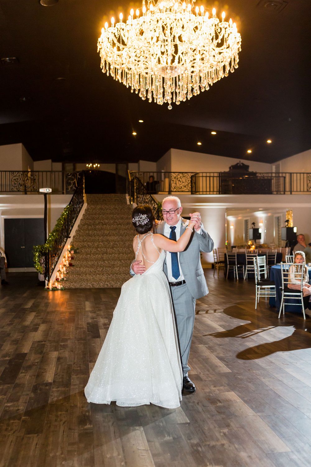 A couple shares a dance under an ornate crystal chandelier in an elegant ballroom venue.