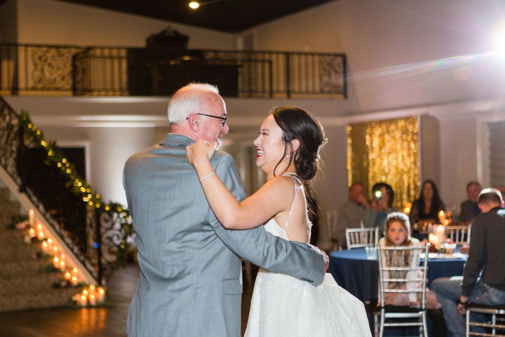 A bride in white dances with a man in grey during an emotional moment at a wedding reception.