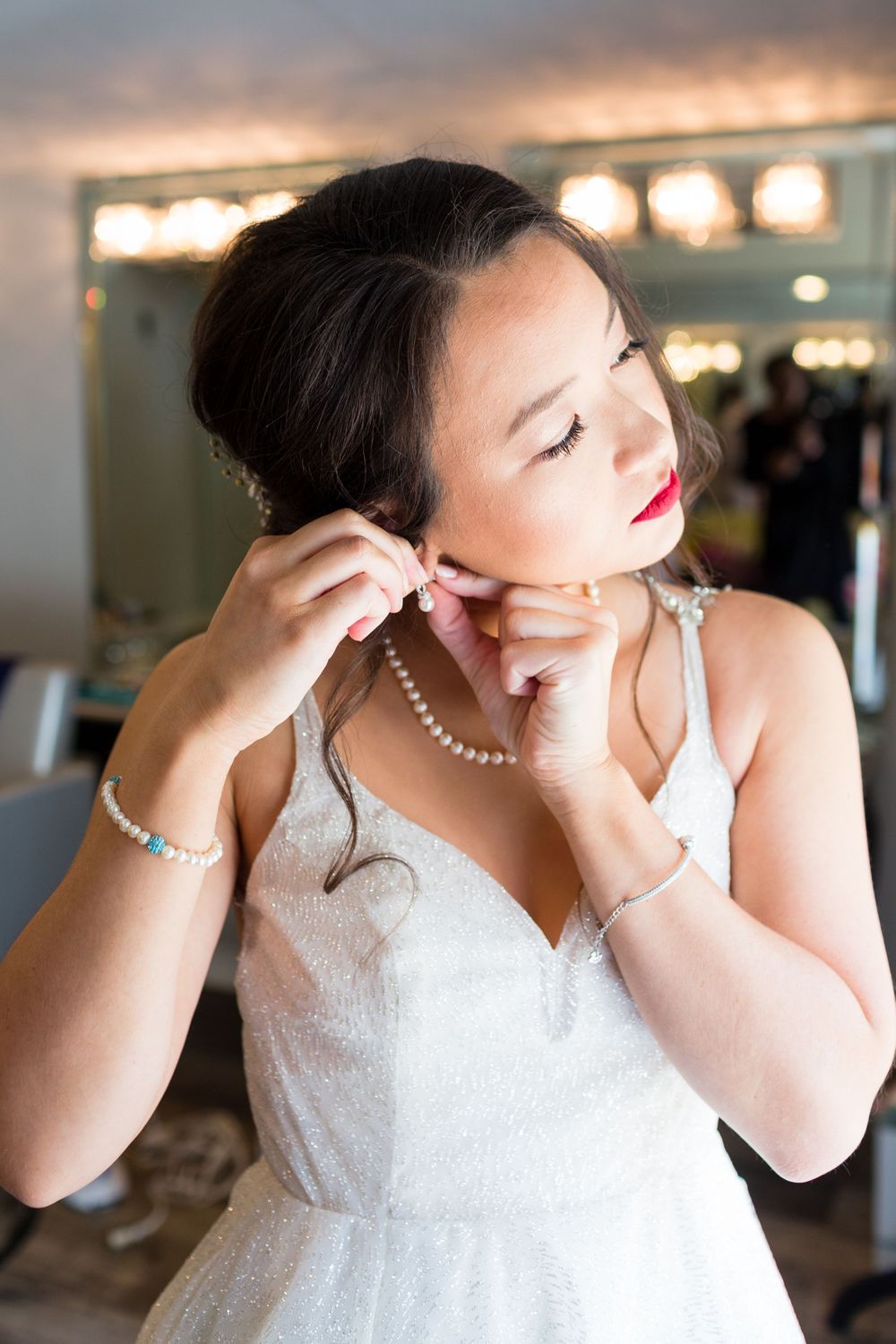 A bride in a white dress puts on pearl jewelry while getting ready in a softly lit dressing room.