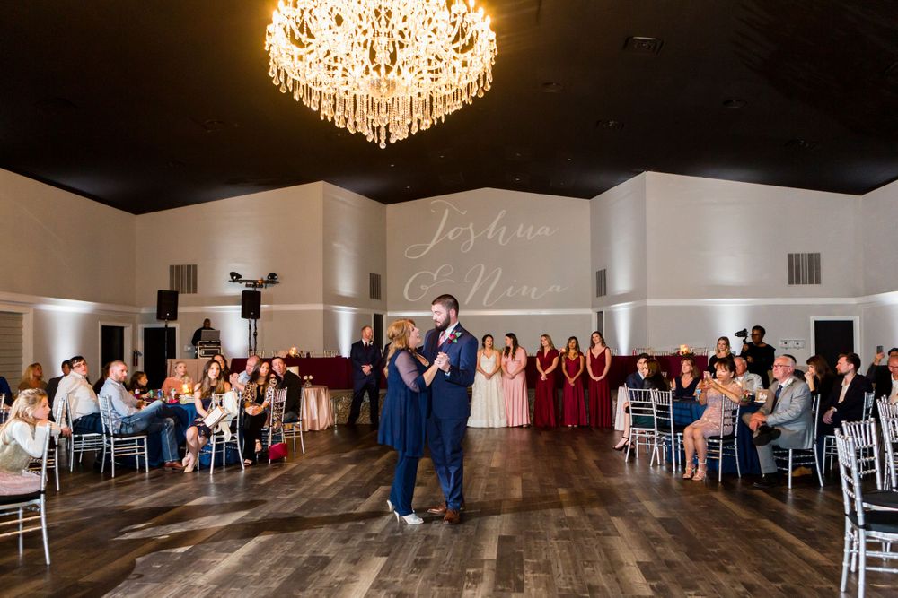 Wedding guests watch a couple dance in an elegant ballroom with a large crystal chandelier overhead.