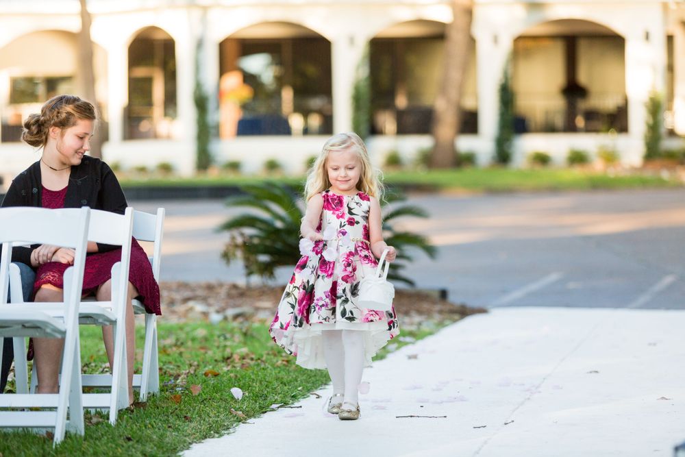 Young flower girl in floral dress walks down outdoor ceremony aisle.