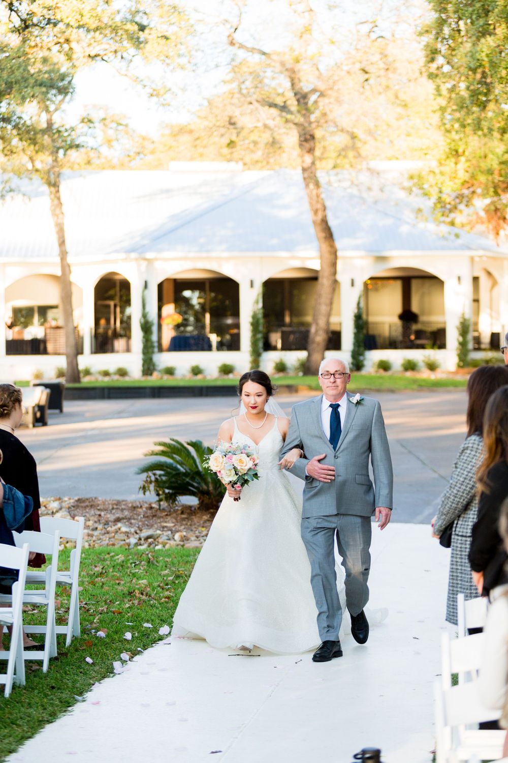 Outdoor wedding processional on white aisle runner with colonial style building in background.