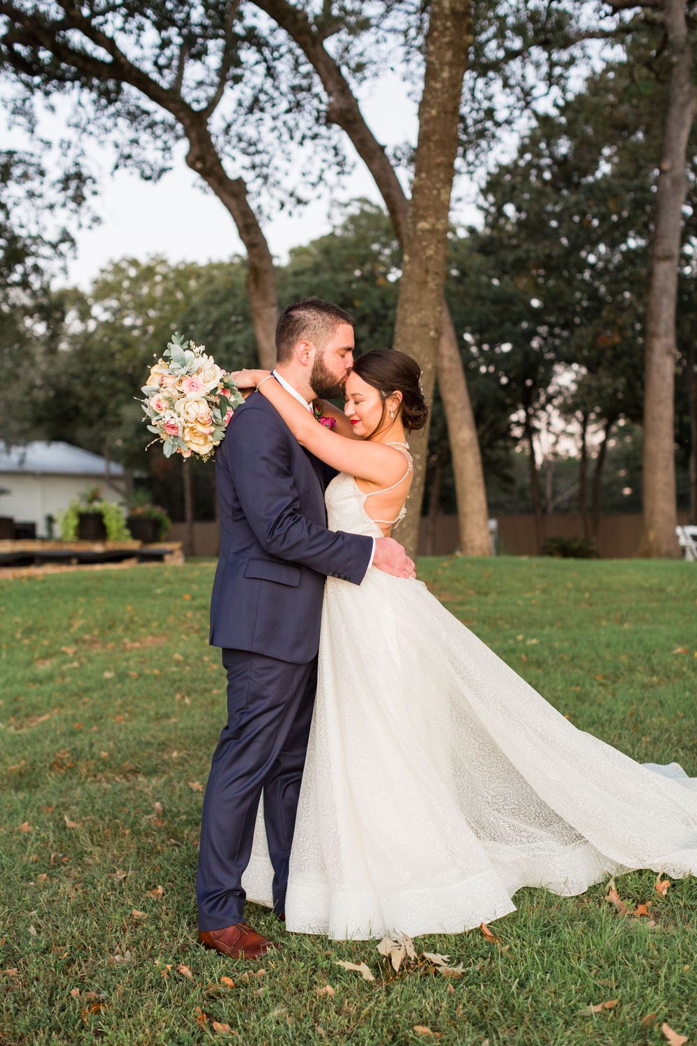 A romantic wedding couple embraces on a grassy lawn with tall trees in the background during golden hour.