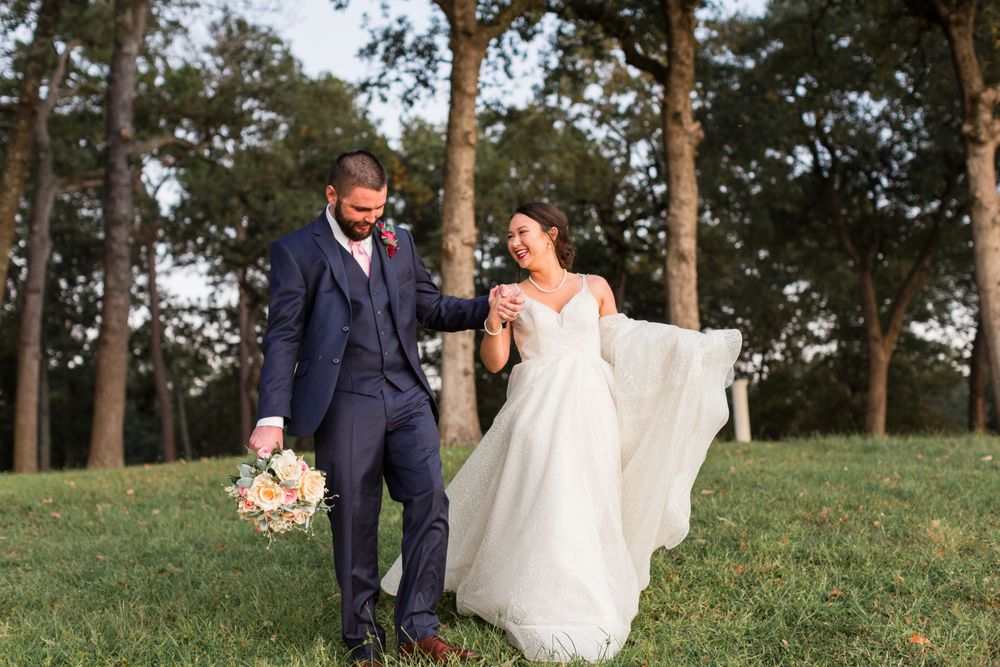 A joyful wedding couple walks together on grass holding a peach bouquet with trees behind them.