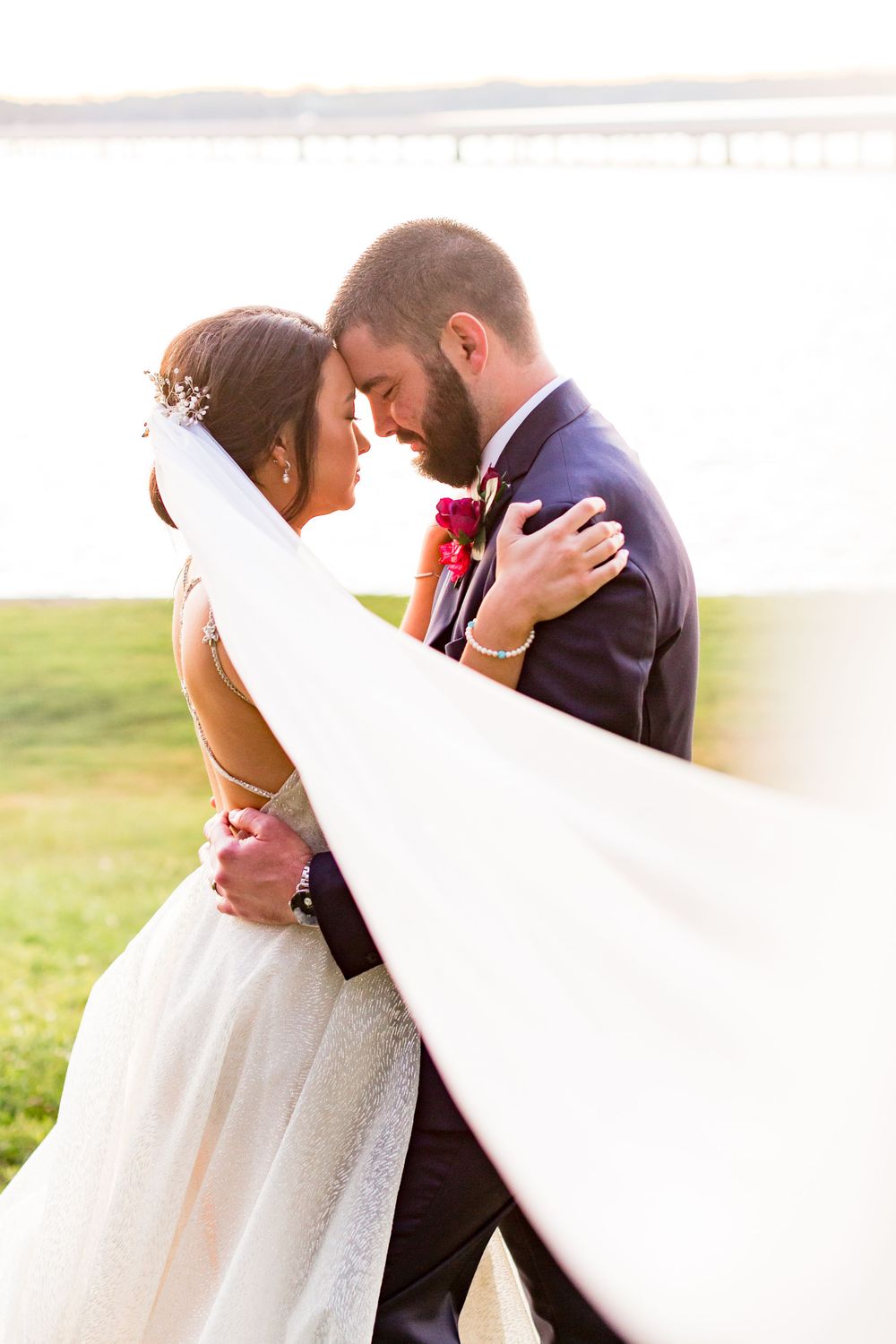 A tender wedding moment captured with flowing white veil against a waterfront backdrop.