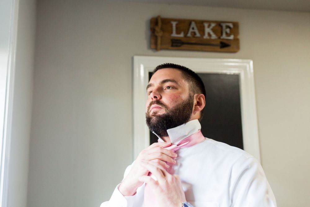A groom in a white shirt adjusts his pink tie while standing beneath a rustic lake sign.