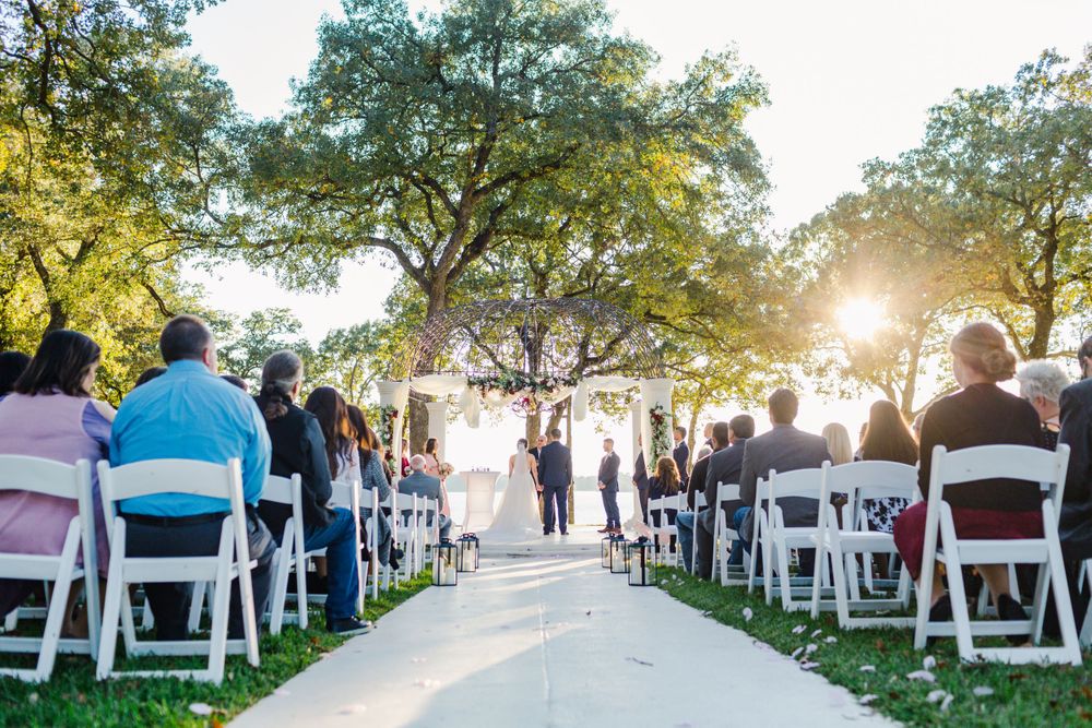 Wedding ceremony setup outdoors with white chairs and trees backlit by golden sunlight.