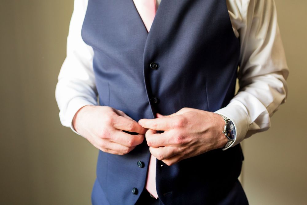 Close-up of hands buttoning a navy blue vest with a pink tie and white shirt.