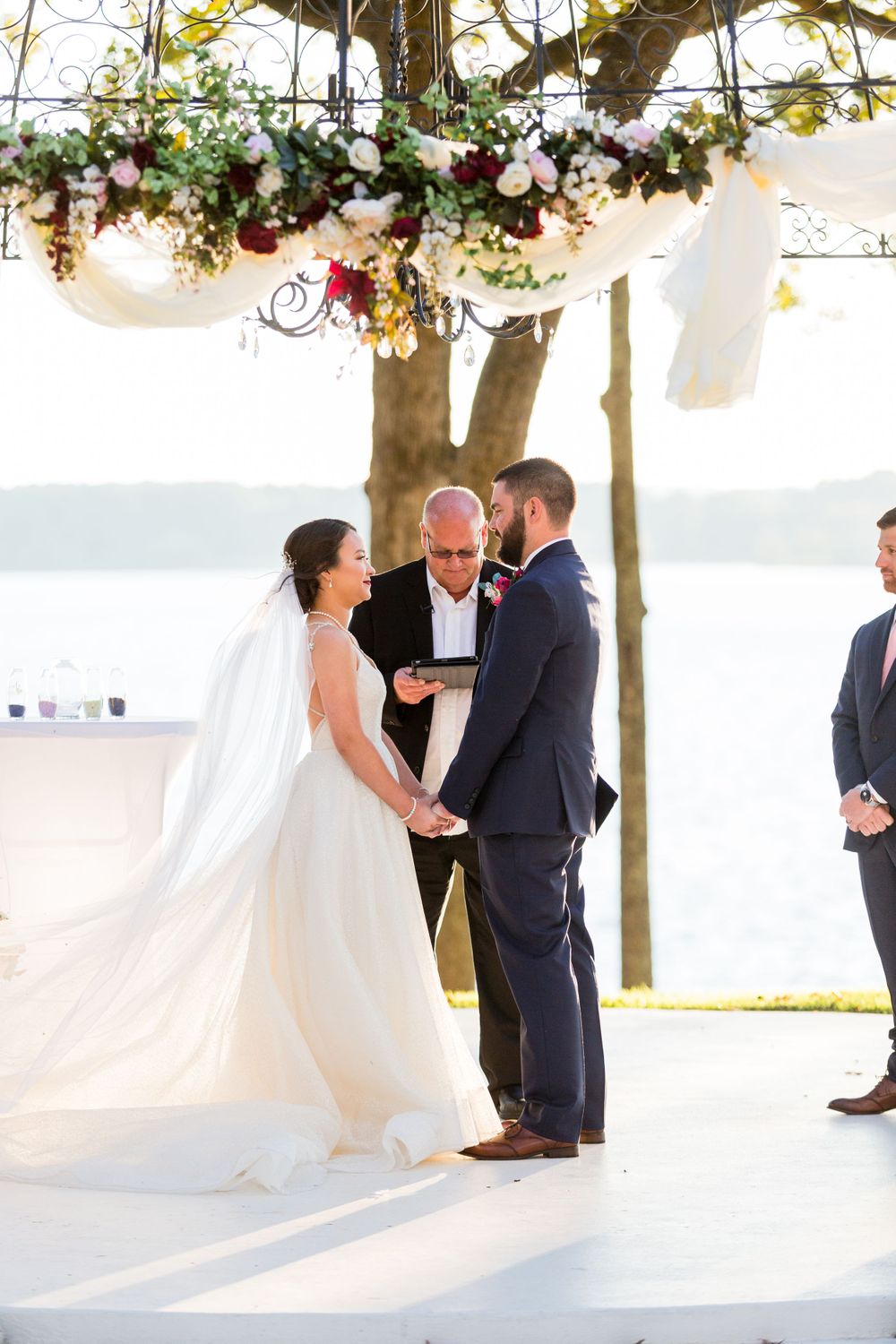 Outdoor lakeside wedding ceremony under floral archway with flowing white fabric and greenery.
