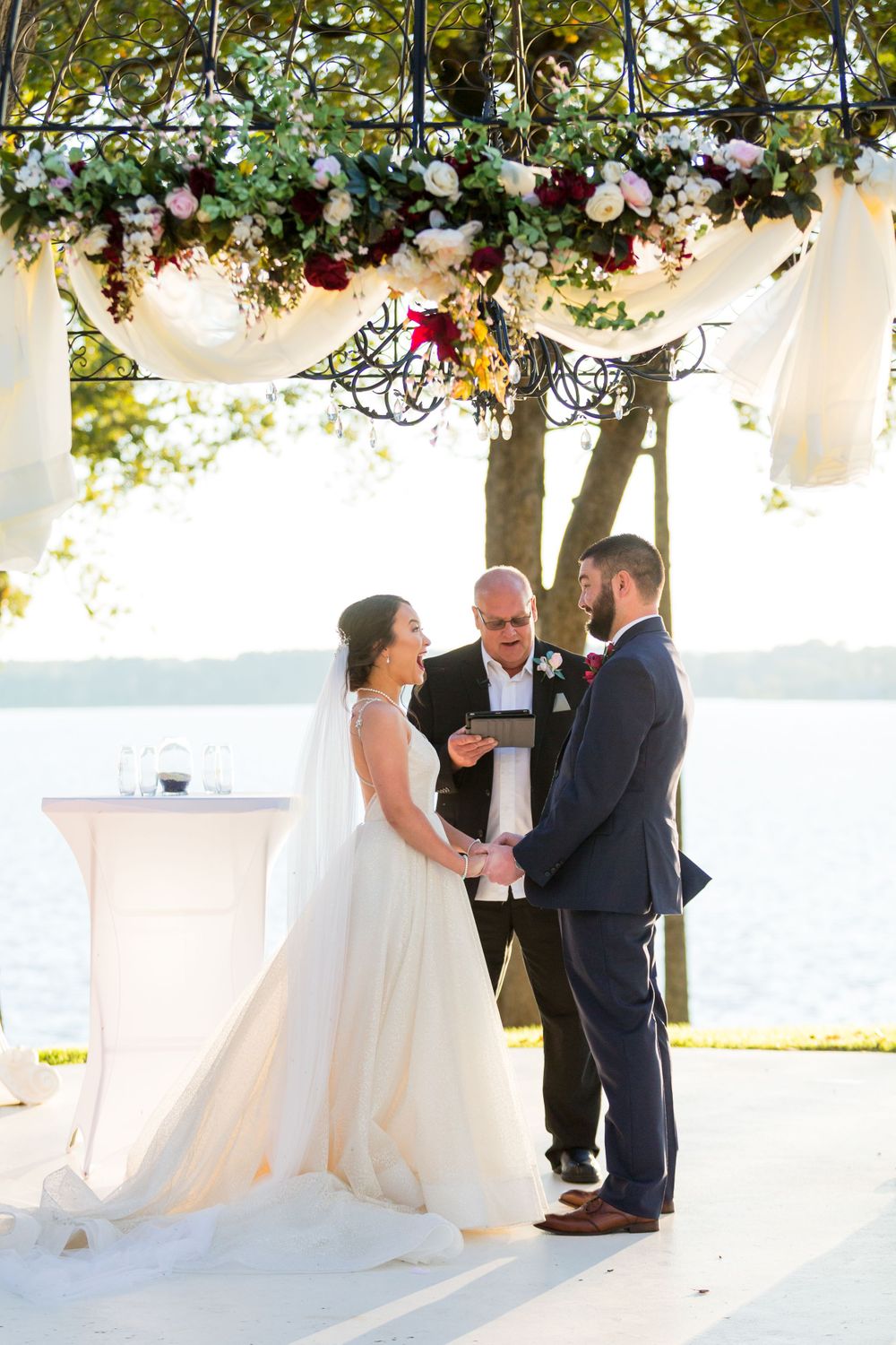 Wedding ceremony by the lake with white floral arrangements and draped fabric decor overhead.