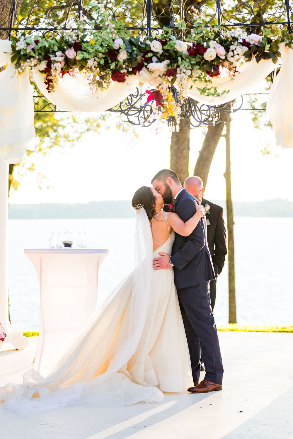 Romantic wedding kiss under a floral-adorned arbor by the waterfront.