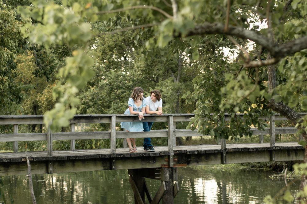 A couple sits together on a wooden bridge over calm water surrounded by trees.