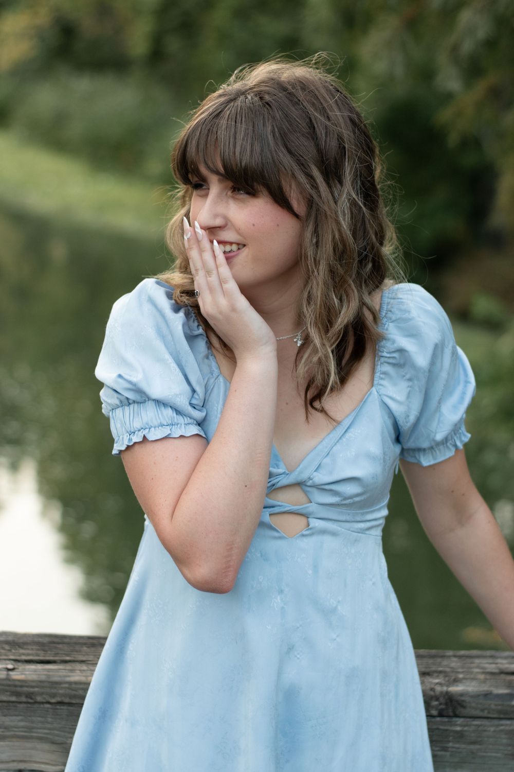A person in a light blue dress with puff sleeves smiling while touching their face.