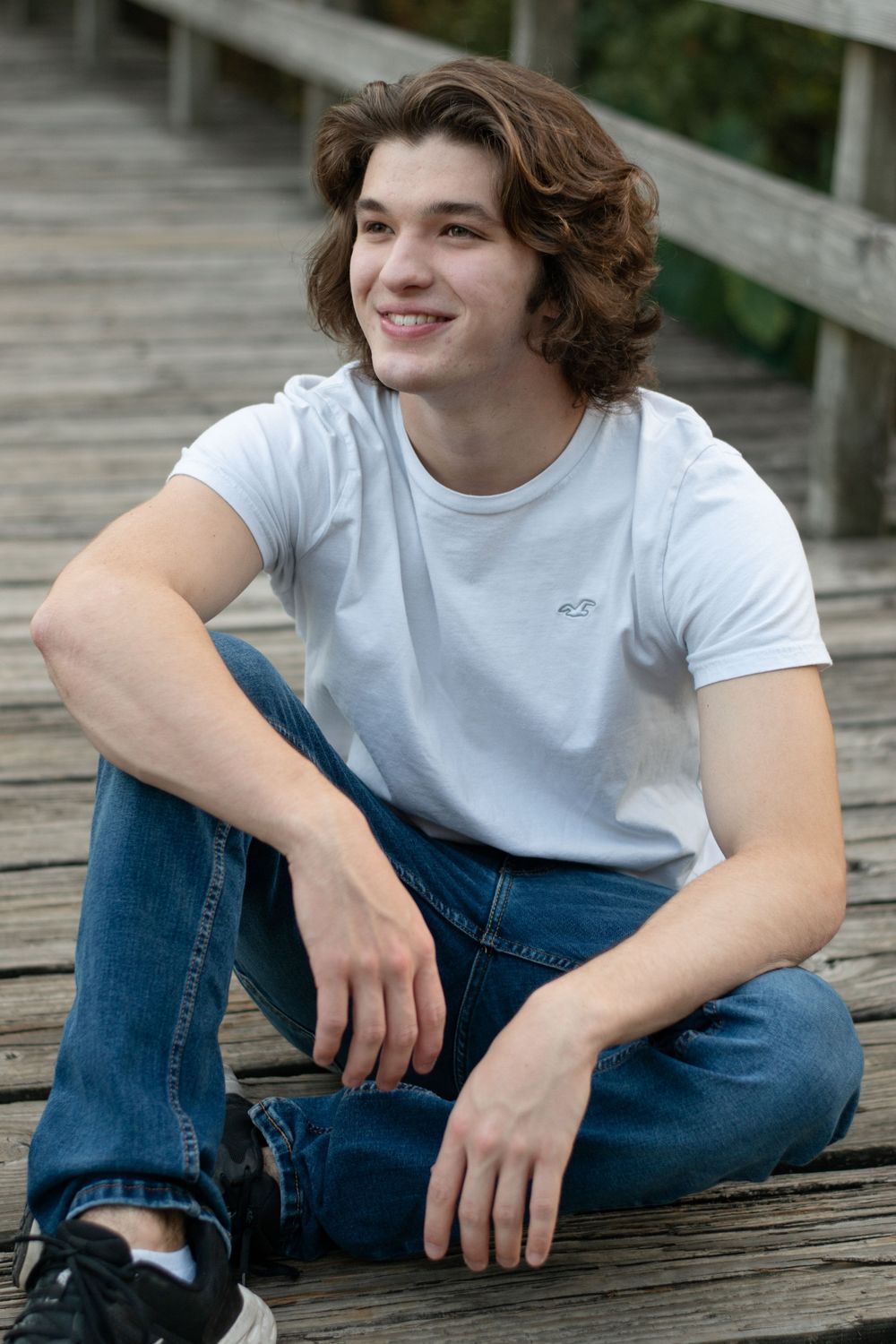 A casual portrait of someone in a white t-shirt and jeans sitting cross-legged on a wooden boardwalk.