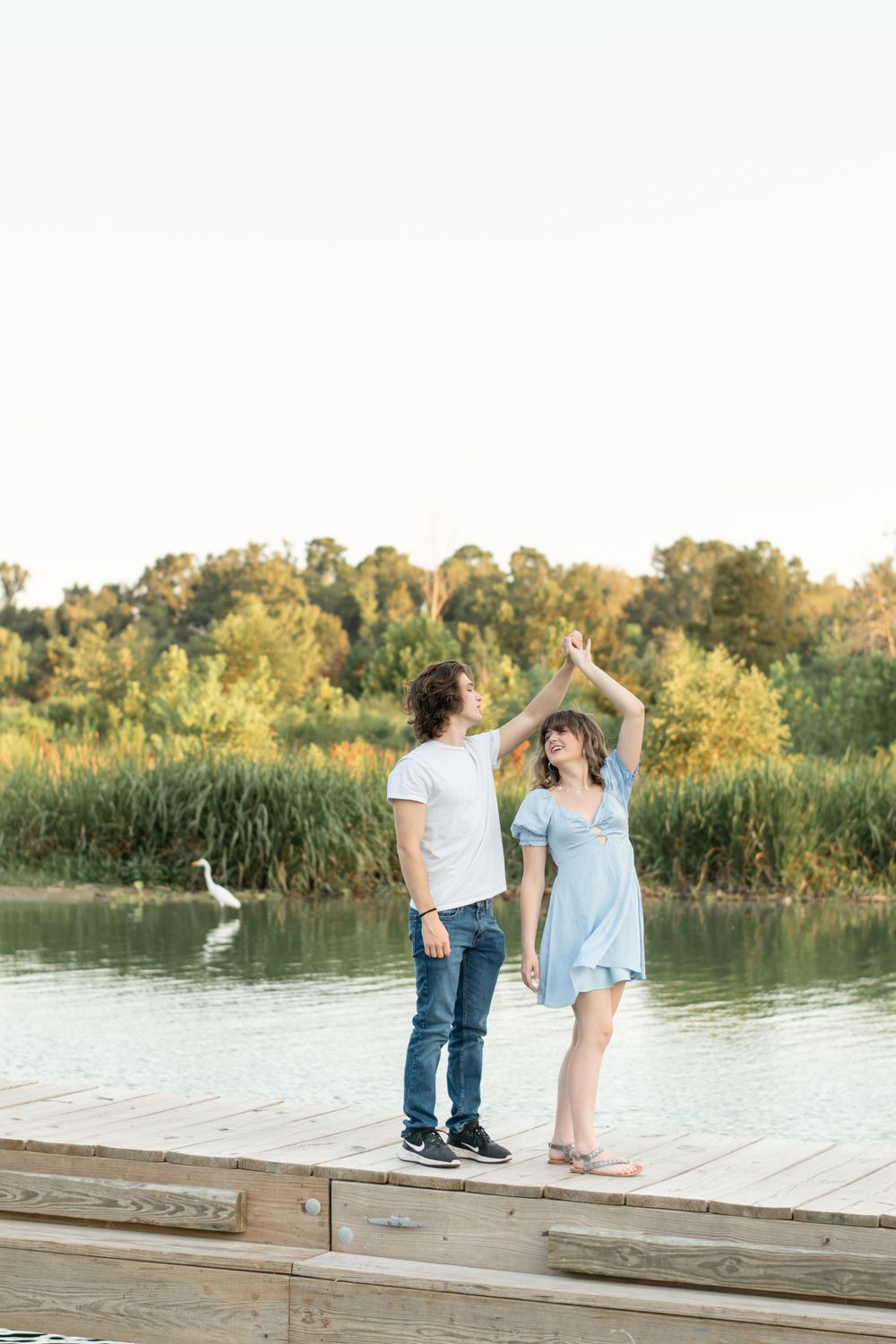 A couple dances together on a dock by a lake with a white egret in the background.