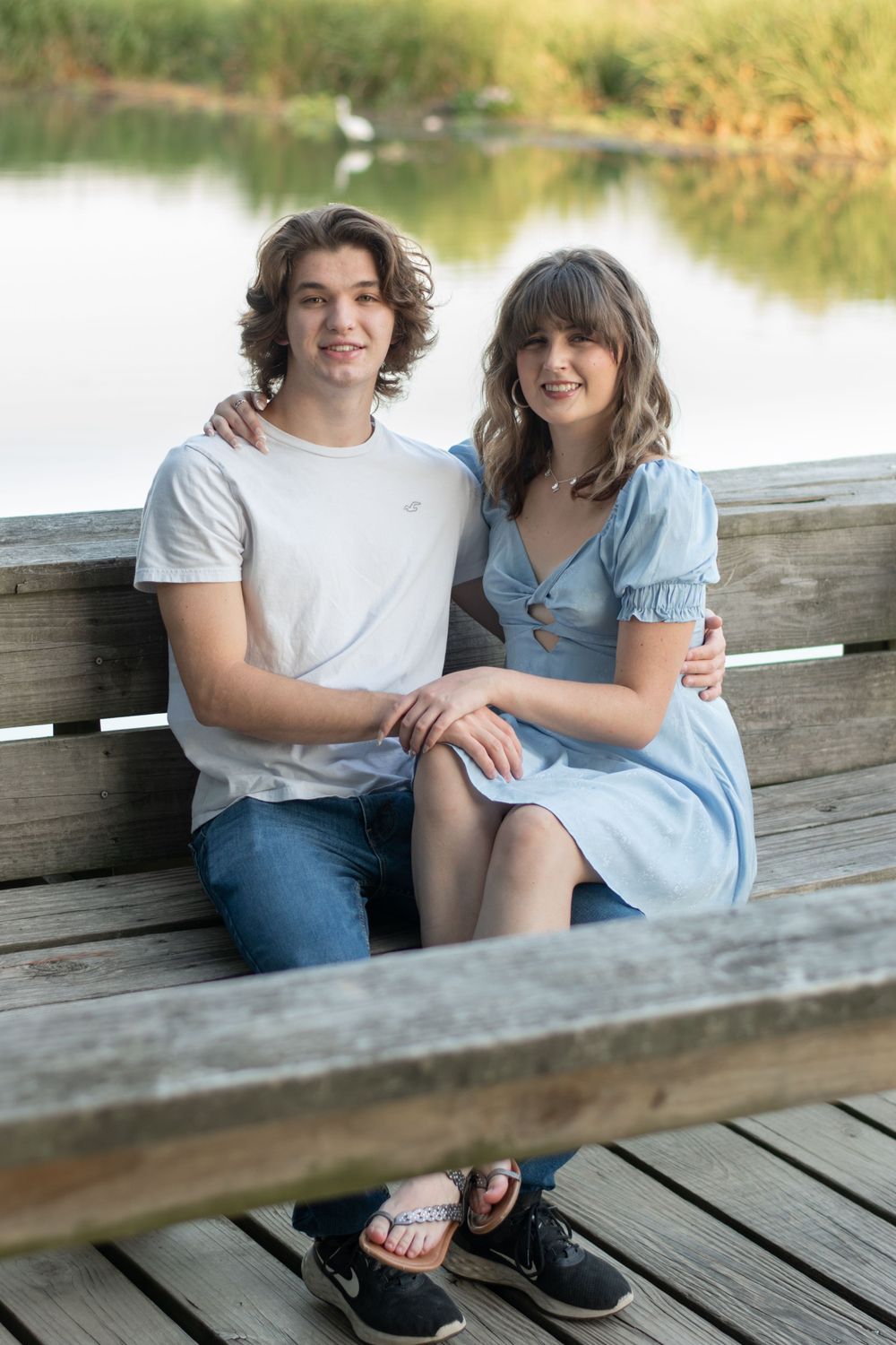 A couple sitting together on a wooden bench by a serene lake in casual summer outfits.