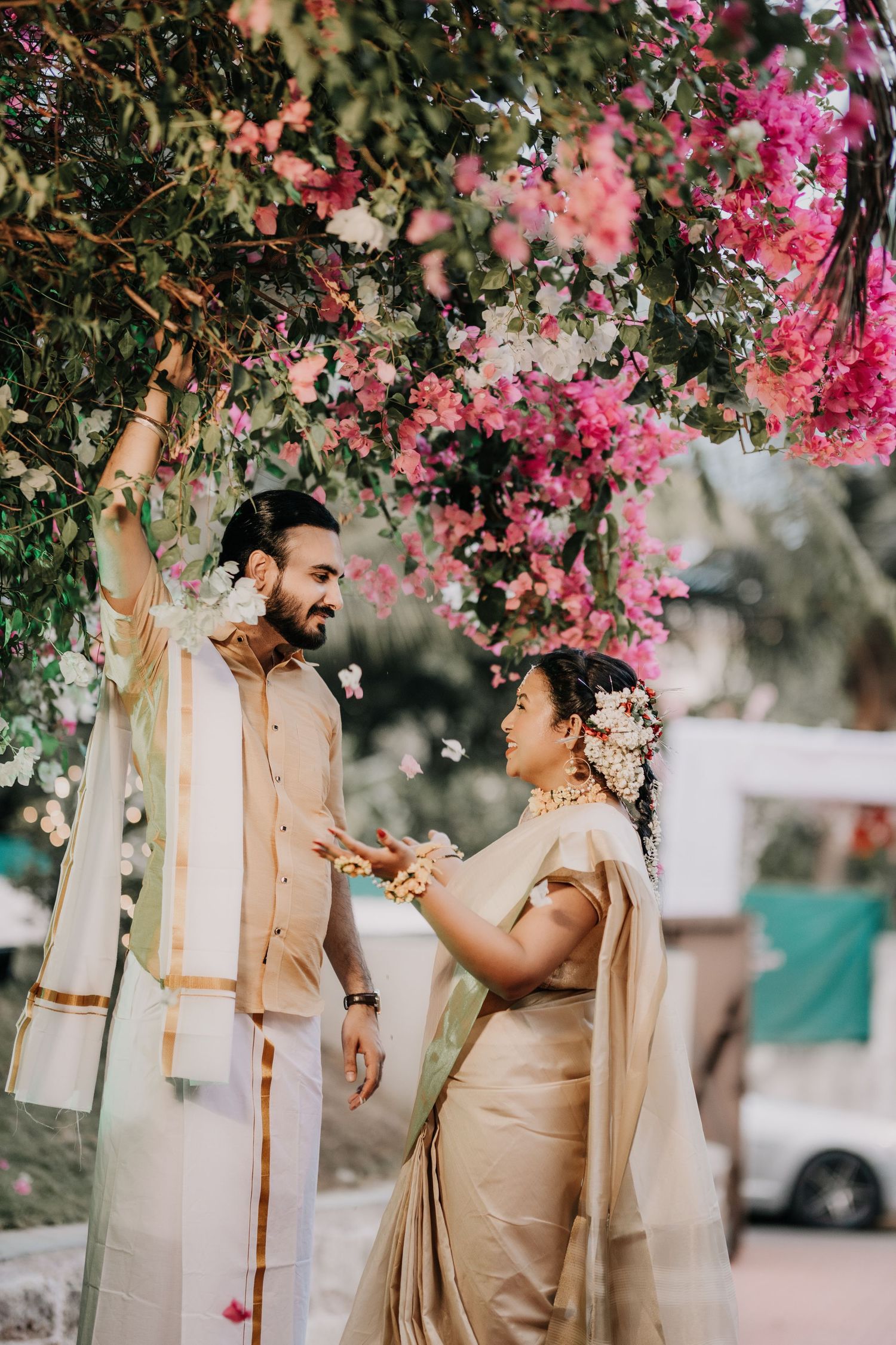 Couple in cream-colored traditional attire shares a joyful moment under pink bougainvillea flowers.