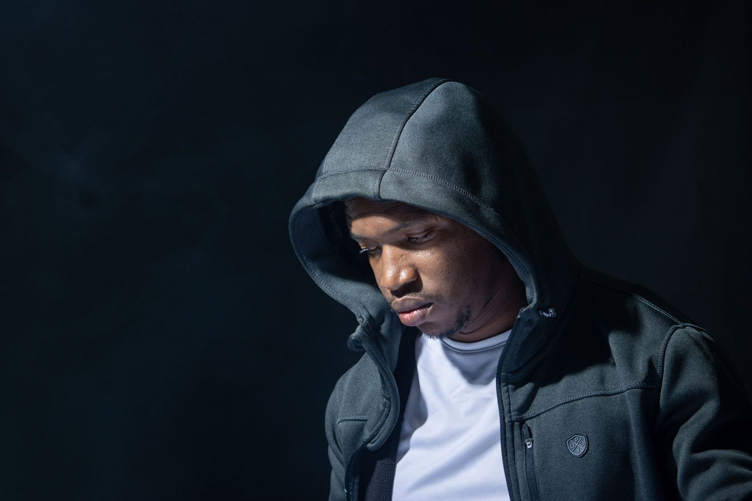 Portrait of person in dark hoodie against black background with dramatic lighting.