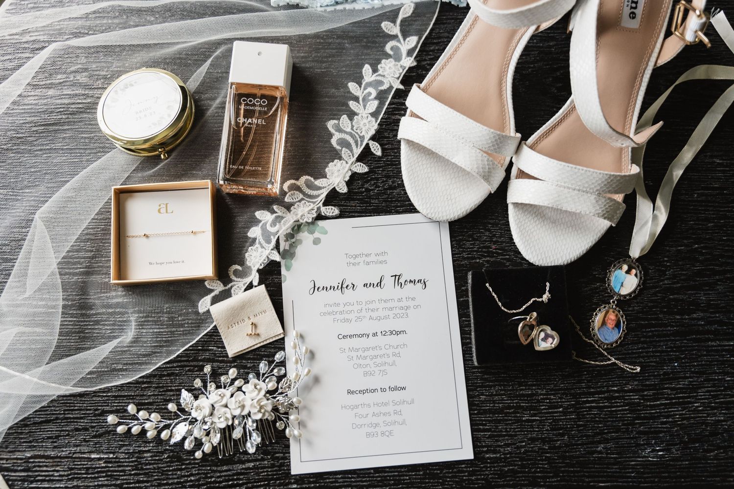 Wedding details including white shoes, invitation, jewelry and floral elements arranged on dark surface.