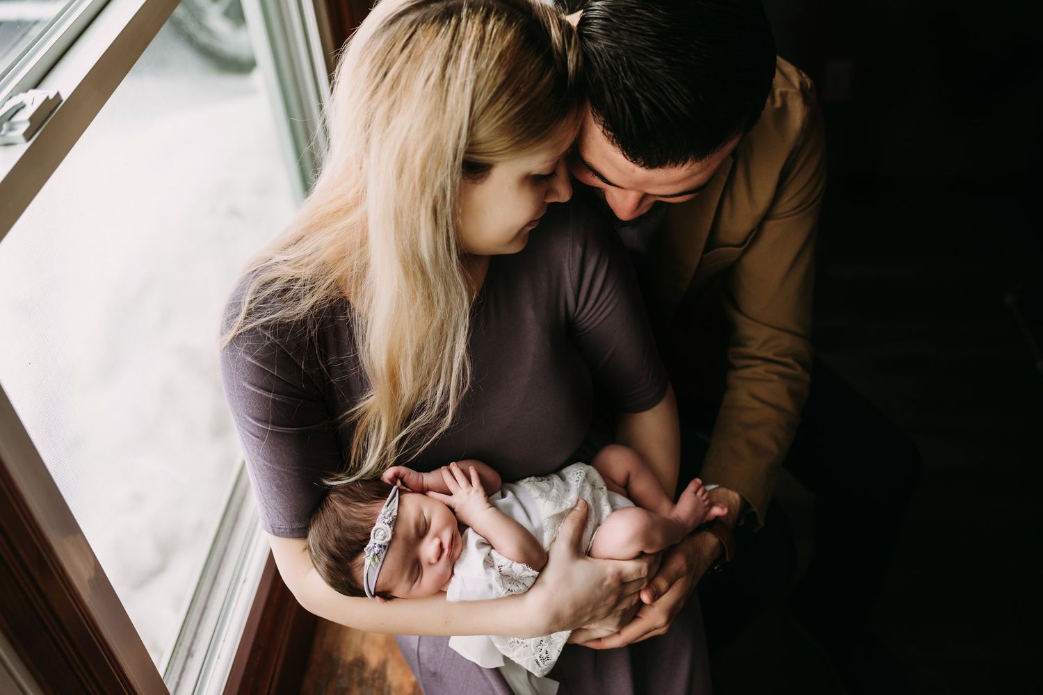 Parents holding newborn baby near window in tender moment.