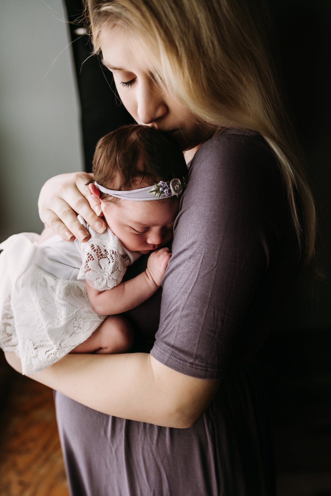 Mother in purple dress tenderly holding sleeping newborn with lace headband.