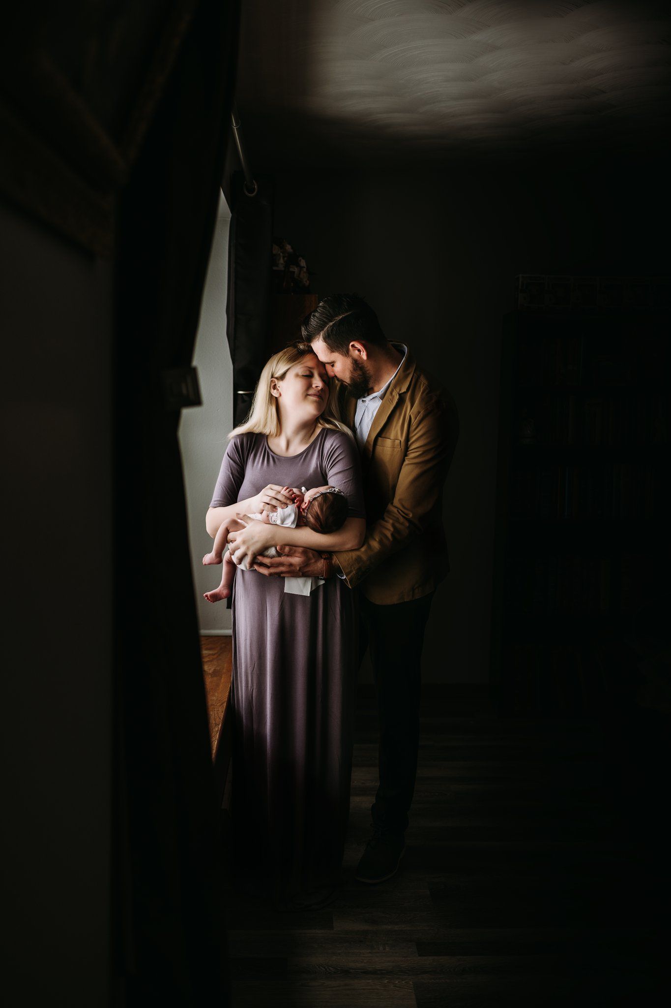 A moody portrait of parents standing in shadow by a window, cradling their newborn.