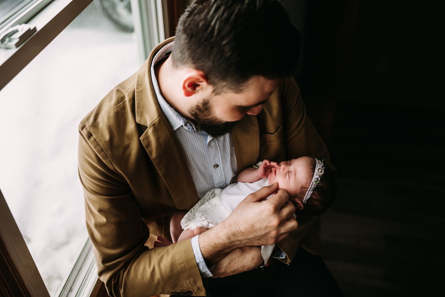 Side profile of someone in a brown blazer tenderly holding a newborn by a window.