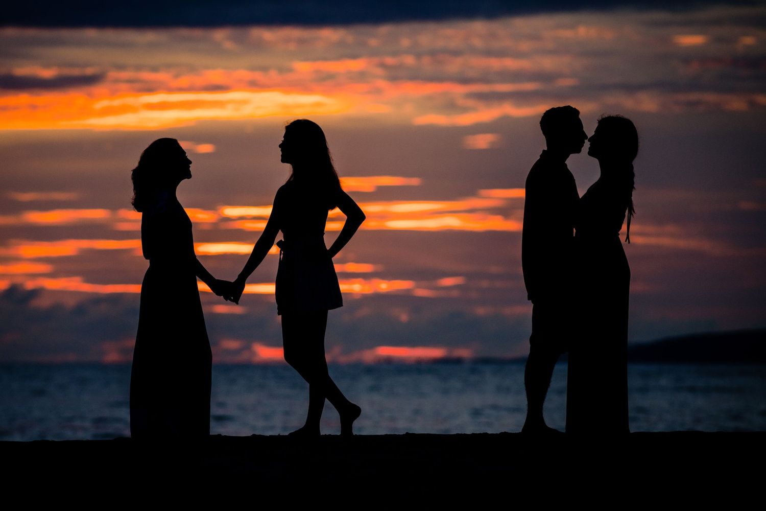 Silhouettes of couples standing on beach against dramatic orange sunset sky.