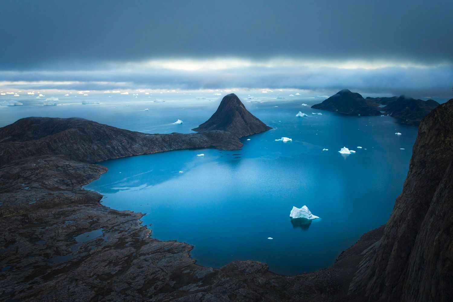 Panoramic view of fjord with icebergs floating in deep blue waters surrounded by mountains.