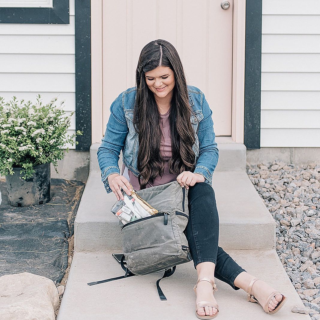 Woman in denim jacket sitting on porch step organizing contents of gray backpack.