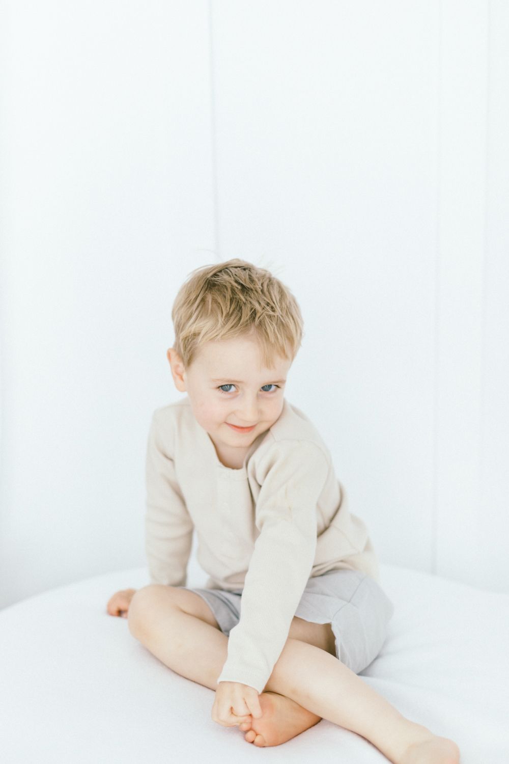 Young child in beige sweater sitting cross-legged on white surface.