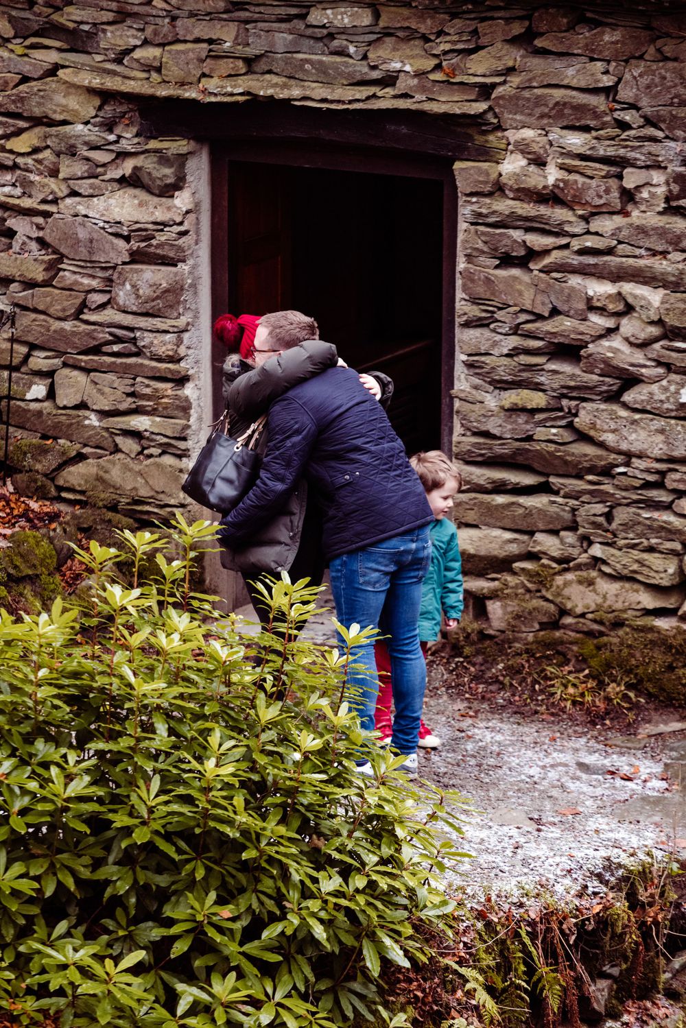 Two people peering through a stone doorway entrance surrounded by rustic stonework and green foliage.