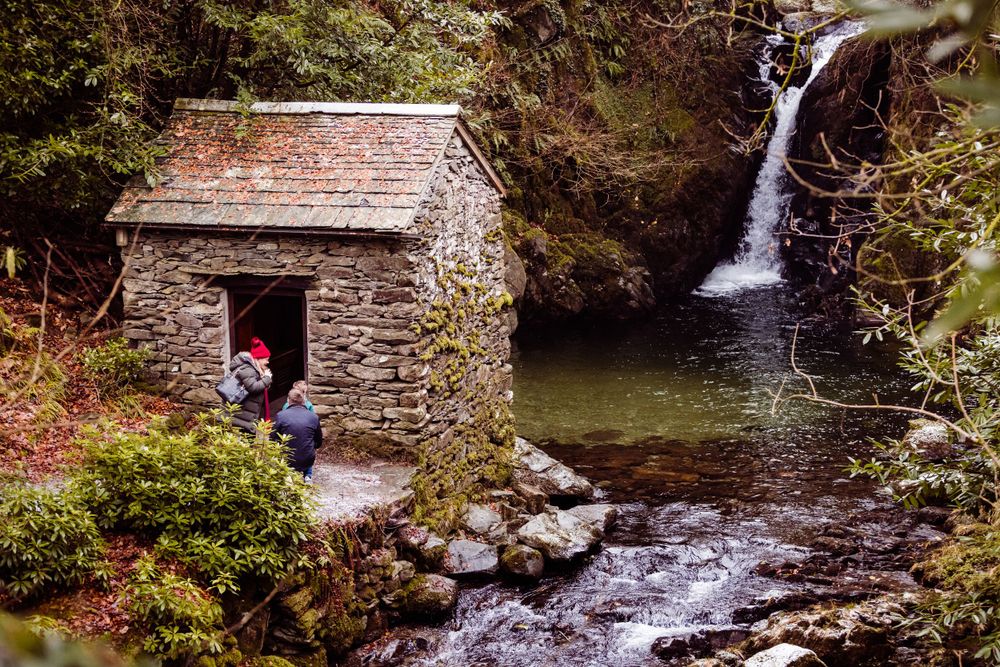 A stone cottage nestled beside a cascading waterfall in a lush woodland setting.