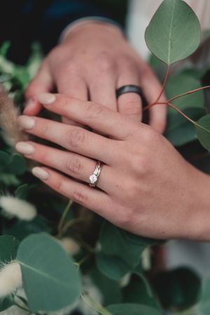 Close up of hands with wedding rings intertwined among green eucalyptus leaves.