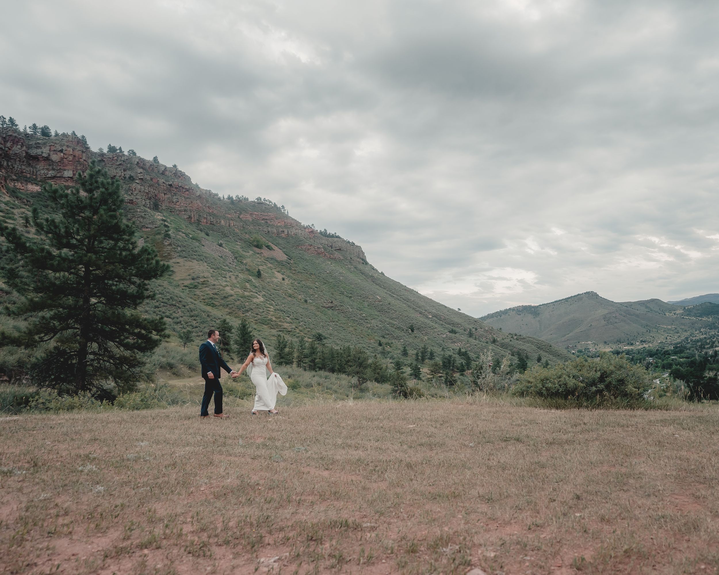 Wide landscape shot of newlyweds in mountain meadow under dramatic sky.