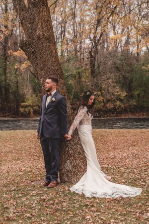 Artistic wedding portrait of couple posed against large tree trunk surrounded by fallen leaves.