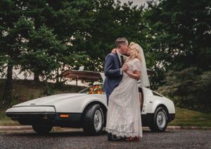 Newlyweds share romantic moment beside classic white sports car.