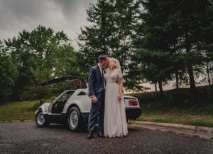 Bride and groom embrace by vintage car at outdoor wedding.