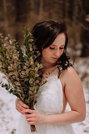 A bride in white dress holding greenery bouquet against snowy winter forest backdrop.