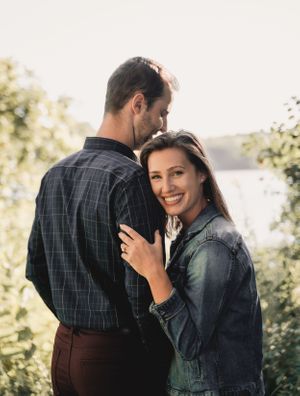 A romantic couple embraces outdoors in casual attire against a sunlit natural backdrop.