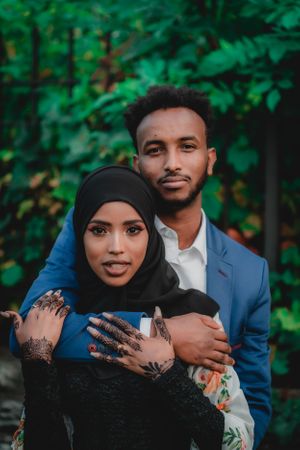 A couple in formal attire poses for a romantic portrait against lush greenery.