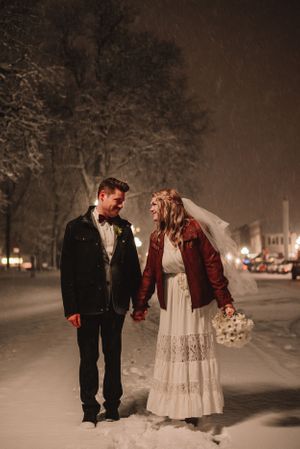 A winter wedding couple in formal attire walking hand in hand through snow covered streets at night.
