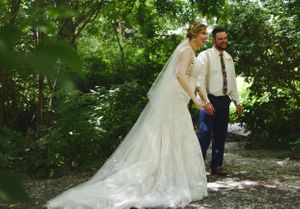 A couple walks along a wooded path on their wedding day with a flowing lace wedding gown trailing behind.