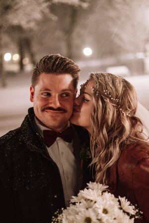 A romantic winter wedding portrait showing a sweet kiss with snow falling and daisies in hand.