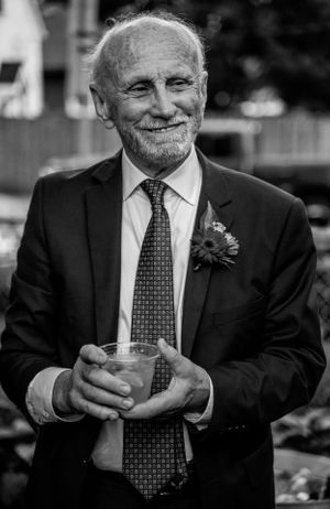Smiling elderly gentleman in formal attire with boutonniere holding a drink at an outdoor event.