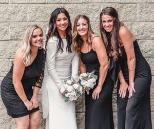 A bridal party in black dresses poses with a bride in white against a stone wall.