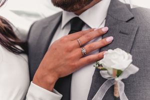 Close-up of bride's hand with engagement ring resting on groom's grey suit lapel with white boutonniere.