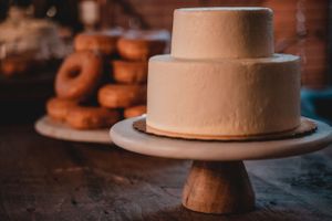 Simple two-tiered beige wedding cake on wooden cake stand with donuts in background.