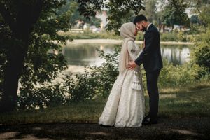 A couple in wedding attire embraces by a serene pond surrounded by trees.